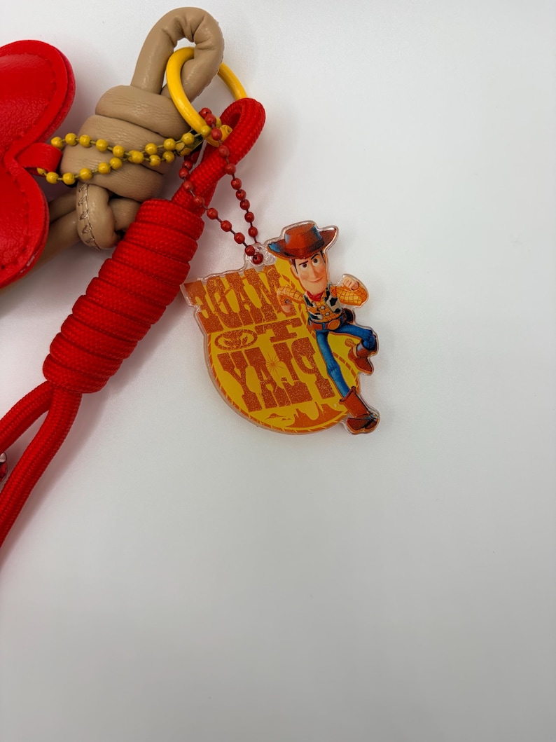 Pu&ograve; includere: Un portachiavi con un cordino intrecciato rosso, una corda annodata color marrone chiaro e un ciondolo a forma di cuore rosso. &Egrave; presente un pendente in acrilico trasparente che raffigura il personaggio Woody di Toy Story con la scritta "TOY STORY" in giallo.