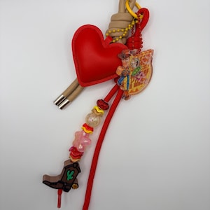 Pu&ograve; includere: Un portachiavi decorativo con un ciondolo a forma di cuore rosso, un cappio color marrone chiaro e un cordino rosso. Include un ciondolo in acrilico trasparente con un personaggio dei cartoni animati, uno stivale da cowboy marrone e perline colorate.