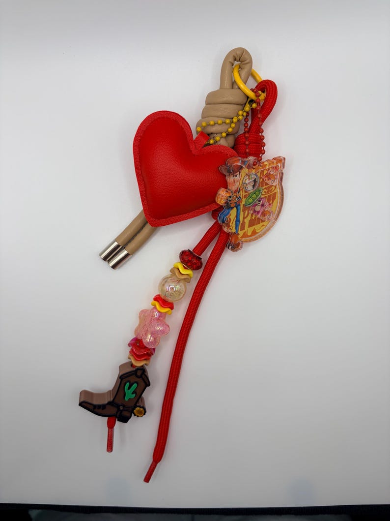 Pu&ograve; includere: Un portachiavi decorativo con un ciondolo a forma di cuore rosso, un cappio color marrone chiaro e un cordino rosso. Include un ciondolo in acrilico trasparente con un personaggio dei cartoni animati, uno stivale da cowboy marrone e perline colorate.