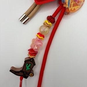 Pu&ograve; includere: Un ciondolo decorativo con uno stivale da cowboy marrone con un dettaglio a cactus verde, infilato su un cordino rosso con perline assortite tra cui una stella rosa, dischi ondulati gialli e rossi e una sfera trasparente.