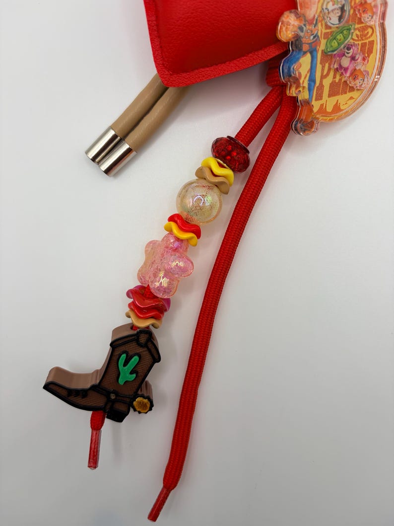 Pu&ograve; includere: Un ciondolo decorativo con uno stivale da cowboy marrone con un dettaglio a cactus verde, infilato su un cordino rosso con perline assortite tra cui una stella rosa, dischi ondulati gialli e rossi e una sfera trasparente.
