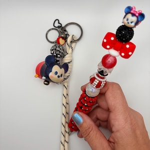 Op de afbeelding: Twee sleutelhangers met Disney-personages. Eén sleutelhanger heeft een Mickey Mouse-bedel en een zwart en zilveren kralenaccent. De andere sleutelhanger heeft een Minnie Mouse-bedel, een rode strik met witte stippen en rode en zilveren kralenaccenten.