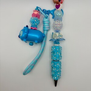 May include: A decorative pen with a blue and pink cord, featuring a Stitch Tsum Tsum charm and various beaded accents. The pen's barrel is adorned with blue flowers and rhinestones, and it has a black tip.