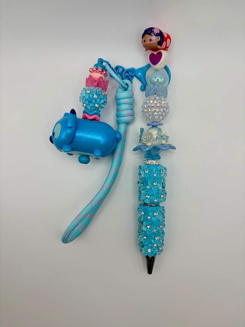 May include: A decorative pen with a blue and pink cord, featuring a Stitch Tsum Tsum charm and various beaded accents. The pen's barrel is adorned with blue flowers and rhinestones, and it has a black tip.