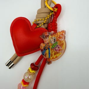 Pu&ograve; includere: Un portachiavi con un ciondolo rosso a forma di cuore, un elemento in corda beige avvolta e un pendente in acrilico trasparente raffigurante un personaggio cowboy. Include un cordino rosso, una catenella gialla e varie perline colorate.