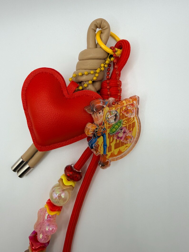 Pu&ograve; includere: Un portachiavi con un ciondolo rosso a forma di cuore, un elemento in corda beige avvolta e un pendente in acrilico trasparente raffigurante un personaggio cowboy. Include un cordino rosso, una catenella gialla e varie perline colorate.