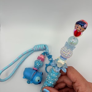 May include: A blue pen with a decorative design and a matching keychain. The pen features a blue barrel adorned with sparkling embellishments and a charm of a character. The keychain has a blue character charm and decorative beads.