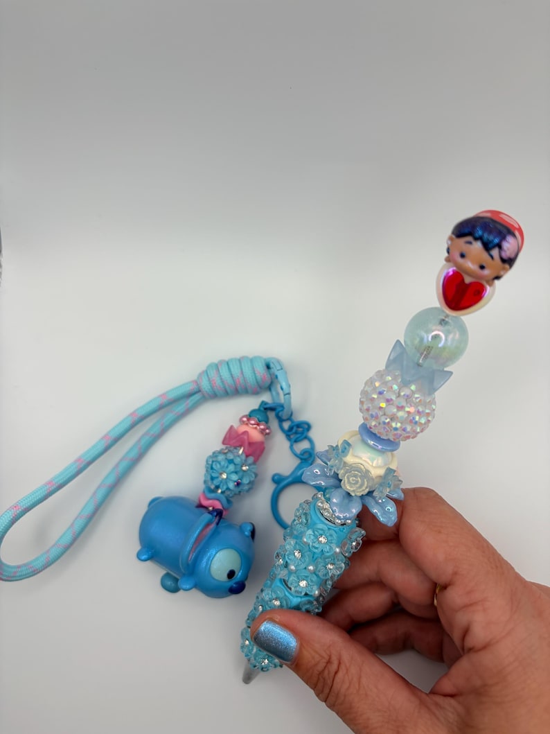 May include: A blue pen with a decorative design and a matching keychain. The pen features a blue barrel adorned with sparkling embellishments and a charm of a character. The keychain has a blue character charm and decorative beads.