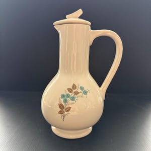 May include: A cream-colored ceramic pitcher with a matching lid and a curved handle. The body of the pitcher features a decorative botanical print consisting of brown leaves and small blue flowers arranged in a cluster on the front surface.