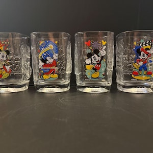 May include: Four clear glass tumblers featuring colorful Mickey Mouse graphics and the year 2000 embossed at the base. Each glass displays a different scene with Mickey Mouse in various outfits, including a wizard robe and director attire, with fireworks.