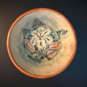 May include: A round ceramic bowl with a terracotta-colored rim and a cream-colored interior. The center features a hand-painted floral design in shades of white, muted green, and soft pink, surrounded by a textured, weathered blue-grey wash.