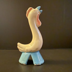 May include: A ceramic rooster figurine in cream, pink, and blue. The rooster has a long, curved neck, open beak, and a stylized base. The figurine is a decorative item, likely for display.