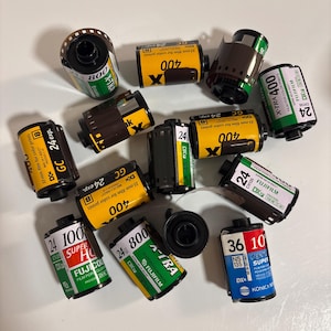 May include: A collection of twelve 35mm film canisters, including various Fujifilm X-Tra 400, 800, and Superia 100, 400, and 800 rolls, alongside a Konica Centuria Super 100 roll. The canisters feature yellow, green, and blue labels with text.