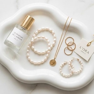 Peut inclure: Un plateau en céramique blanche contenant un flacon de parfum en verre avec un bouchon doré étiqueté LE PARFUM DE L'AMOUR, deux bracelets de perles, un collier à chaîne dorée avec un pendentif rond, trois bagues dorées, des créoles en perles.