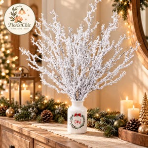 May include: A white ceramic vase featuring a green wreath graphic with a red bow and the text Joy, containing a large arrangement of white, snow-dusted branches that spread outward in a tall, multi-stemmed, tree-like structure.