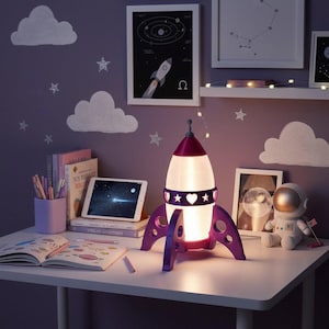 May include: A purple and white rocket ship lamp with heart and star cutouts, sitting on a white desk. The lamp is lit, and the room has a space theme with star and cloud wall decals. A tablet and books are also on the desk.