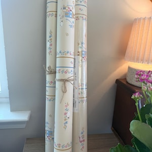 May include: Three rolls of cream-colored wallpaper tied together with twine. The wallpaper features a repeating pattern of small blue houses, sheep, and floral garlands in shades of pink, blue, and green, separated by thin horizontal blue stripes.