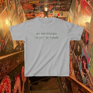 May include: A heather gray short-sleeve t-shirt featuring the text "MY BOYFRIEND IS OUT OF TOWN" printed in a dark gray, sans-serif font across the center of the chest. The garment has a crew neckline and a standard, straight-cut silhouette.