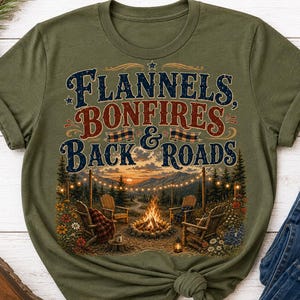 Może przedstawiać: Oliwkowozielony t-shirt z nadrukiem graficznym zawierającym tekst "Flannels, Bonfires & Back Roads" nad ilustracją przedstawiającą ognisko, drewniane krzesła, girlandy świetlne, sosny i góry o zachodzie słońca.
