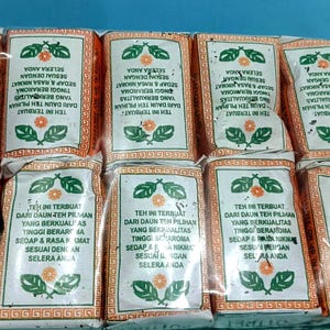 May include: A set of ten rectangular tea packets arranged in two rows of five, wrapped in clear plastic. Each white packet features an orange border, a central orange flower icon, green leaf illustrations, and black Indonesian text.