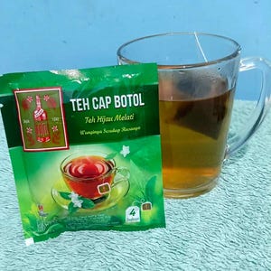 Pode incluir: Um pacote de saquinhos de chá verde com o rótulo Teh Cap Botol Teh Hijau Melati e uma ilustração de uma chávena de chá e uma garrafa vermelha. Ao lado do pacote, uma caneca de vidro transparente cheia de chá âmbar com um saquinho.