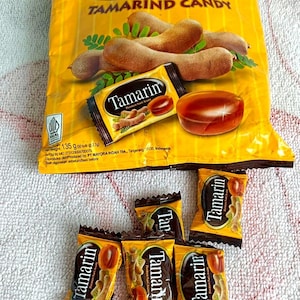 Puede incluir: Un paquete de caramelos de tamarindo con las palabras "Tamarind Candy". Varios caramelos envueltos individualmente están esparcidos alrededor del paquete. El caramelo es de color marrón oscuro.