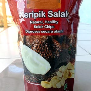Pode incluir: Uma embalagem de plástico vermelha para snacks com o rótulo Keripik Salak, Natural, Healthy Salak Chips, Diproses secara alami. A embalagem apresenta uma imagem de uma fruta-cobra inteira e várias chips fatiadas, com 50 gramas de peso.