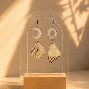 May include: A pair of mismatched earrings featuring silver-toned metal charms. One earring displays a crescent moon above a translucent, veined butterfly wing. The other earring shows a sunburst charm above a similar translucent butterfly wing.