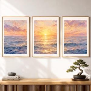 Op de afbeelding: Een drieluik van ingelijste muurkunstprints met een doorlopend zeegezicht bij zonsondergang. De afbeeldingen tonen een gouden zon laag aan de horizon, die licht reflecteert op blauwe oceaangolven onder een lucht vol zachte roze, oranje en paarse wolken.