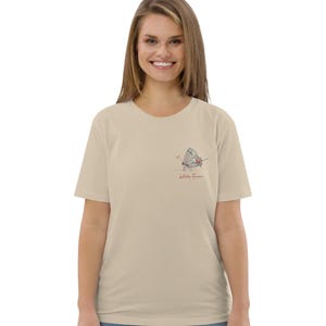 May include: Beige t-shirt featuring a small butterfly illustration with red, blue, and gray accents. The text "Wild as Free" is printed below the butterfly. The shirt is made of a soft material.
