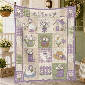 May include: A quilted blanket featuring a grid of squares with illustrations of ducks, lavender bouquets, a house, a watering can, a basket, a heart, a hat, and boots. The name Olivia is printed at the top in a purple script font.