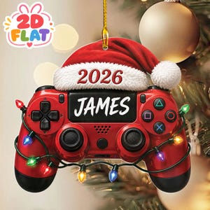 May include: A red video game controller ornament featuring a white-trimmed red Santa hat on top. The text 2026 is printed on the white hat trim, and the name JAMES is printed in white on the centre of the controller. A string of colourful Christmas lights wraps around the controller.