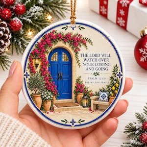 Puede incluir: Un adorno redondo de cerámica con una puerta doble azul rodeada de flores rosas, un farol y un limonero en maceta. El texto dice: THE LORD WILL WATCH OVER YOUR COMING AND GOING, PSALM 121:8, THE WILSON FAMILY, con un cordón dorado para colgar.