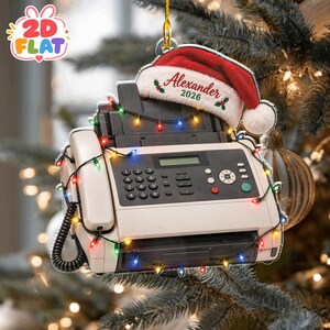 May include: A beige office fax machine ornament wrapped in a string of multicoloured Christmas lights. A red and white Santa hat sits on top of the machine with the text Alexander 2026 embroidered in red script with small green holly leaves.