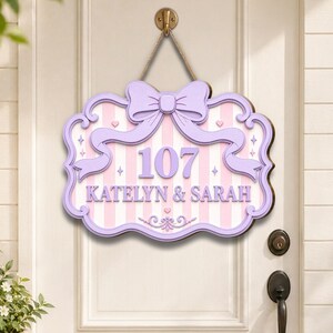 May include: A decorative wall sign featuring a light purple bow at the top, a pink and white striped background, and the text 107 KATELYN & SARAH in purple lettering. The sign has a scalloped border and is suspended by a rope from a metal hook.