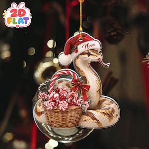 May include: A flat hanging ornament featuring a tan and brown snake wearing a red and white Santa hat with the name Nova printed on the brim. The snake is coiled around a woven brown basket filled with red and white striped candy canes.