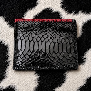 May include: A rectangular black wallet featuring a textured snakeskin pattern across the surface. The top edge is accented with a distinct row of bright red decorative stitching that contrasts against the dark, glossy scales of the material.