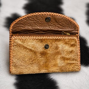 May include: A rectangular clutch wallet featuring a tan, textured fur front panel and a dark brown, pebbled leather flap. The edges are finished with a braided leather trim. It includes a metal snap closure and a top zipper compartment.