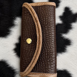 May include: A rectangular dark brown key case with a textured crocodile-embossed pattern. The case features a curved flap secured by a single polished gold-tone metal snap button and is finished with light tan leather piping along the edges.
