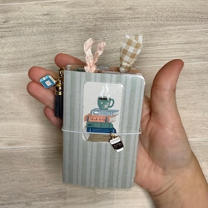 May include: A small, light green striped notebook featuring a central illustration of a stack of books topped with a steaming mug. The notebook is accented with a pink ribbon, a beige gingham ribbon, a blue book charm, a tassel, and a coffee cup charm.