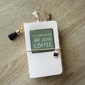 May include: A small notebook with a light pink cover featuring a repeating cross pattern. A green rectangular label on the front reads, "SIT BACK, RELAX and drink COFFEE." It includes a black tassel, a coffee cup charm, and two fabric ribbons.