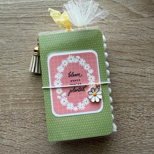 May include: A small green notebook with a polka dot pattern, featuring a pink rectangular label with a white daisy wreath and the text "bloom where you're planted." It includes a white elastic band, a daisy charm, a tassel, and white tulle trim.