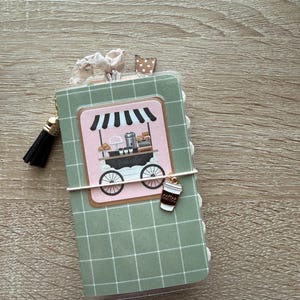 May include: A small notebook with a sage green and white grid cover featuring a central illustration of a coffee cart under a striped awning. The notebook is accented with a black tassel, a brown polka dot tab, and a small coffee cup charm.