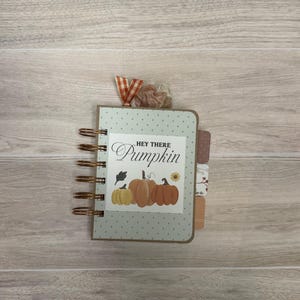 May include: A small spiral-bound notebook with a light green cover featuring a white polka dot pattern. The center label displays the text "HEY THERE Pumpkin" above an illustration of three pumpkins and a sunflower, accented by fabric ribbons.