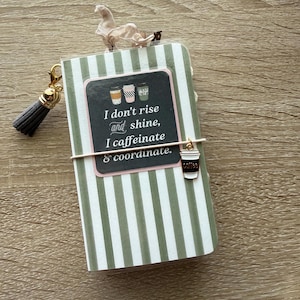 May include: A small notebook with a green and white vertical striped cover featuring a central label that reads, "I don't rise and shine, I caffeinate & coordinate." The notebook includes a grey tassel charm and a small coffee cup charm.