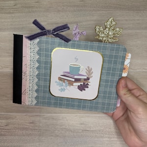 May include: A small handmade journal with a blue plaid cover, a black spine, and a white lace trim. The front features a square illustration of a coffee mug resting on a stack of books, accented by a dark purple velvet bow and a gold glitter leaf.