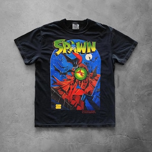 Puede incluir: Una camiseta negra de manga corta con un estampado gráfico del personaje de cómic Spawn. El diseño muestra una figura con capa roja sobre un cielo nocturno azul con luna llena, murciélagos y un orbe verde brillante. La palabra SPAWN está en amarillo.