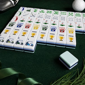May include: A set of white rectangular mahjong tiles featuring golf-themed graphics, including golf clubs, golf carts, trophies, and numbered flags. Each tile has a smooth finish with colorful printed designs and a blue and white layered base.