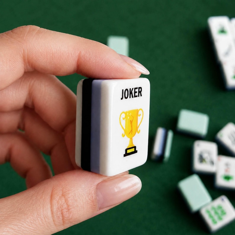 May include: A white rectangular mahjong tile featuring the word JOKER in black text above a yellow trophy icon. The side of the tile displays vertical stripes in black, light purple, and white, with other tiles scattered in the background.