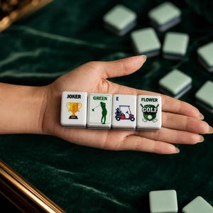 May include: Four rectangular white mahjong tiles featuring golf-themed graphics. The tiles display the text JOKER with a gold trophy, GREEN with a silhouette of a golfer, E with a golf cart, and FLOWER GOLF with crossed golf clubs and a ball.